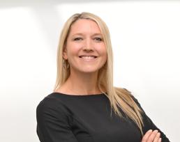 A white woman with long blonde hair and a big smile. Wearing a grey top and hoop earrings. 